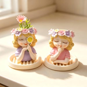 May include: Two decorative resin planters shaped like young figures with blonde hair, wearing floral crowns of pink, purple, and white blossoms. One figure holds a yellow flower, while the other holds a finger to their lips in a hush gesture.