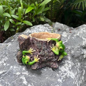 May include: A brown tree stump planter featuring two bright green frogs with large yellow eyes clinging to the sides. The textured bark exterior has a hollow centre for planting, and the frogs are positioned on the front surface of the stump.