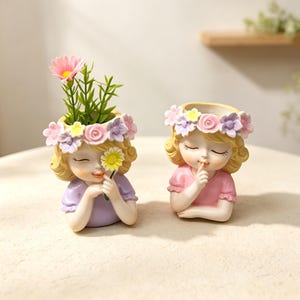 Op de afbeelding: Twee hars plantenbakjes in de vorm van jonge kinderen met blond haar en bloemenkransen. Het linker bakje draagt een paars shirt en houdt een geel madeliefje vast, het rechter bakje draagt een roze shirt en houdt een vinger voor de lippen.