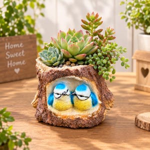 Peut inclure: Un pot de fleurs décoratif en forme de souche d'arbre avec un centre creux contenant deux oiseaux sculptés bleus et jaunes. Le dessus de la souche est rempli d'une variété de plantes succulentes vertes aux pointes roses et feuilles texturées.