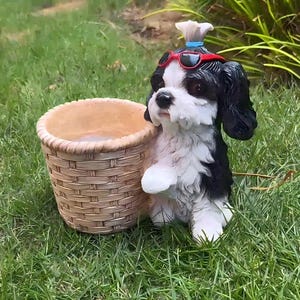 Puede incluir: Una figura de perro en blanco y negro con un peinado de moño y gafas de sol rojas, situada junto a una maceta cilíndrica de color tostado con textura trenzada. El perro tiene orejas oscuras, cara blanca y una pata sobre la maceta.