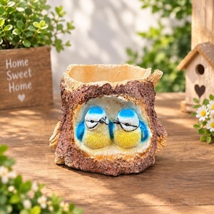 Peut inclure: Un pot de fleurs en forme de souche d'arbre creuse avec un extérieur en écorce brune texturée et une ouverture sculptée à l'avant. À l'intérieur, deux petits oiseaux bleus et jaunes au visage blanc et aux marques oculaires noires sont nichés.