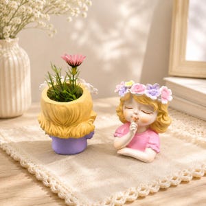 Könnte beinhalten: Zwei Pflanzgefäße aus Kunstharz in Form von Kinderbüsten mit blondem Haar. Ein Gefäß hat eine violette Basis und enthält eine grüne Pflanze mit einer rosa Blüte. Das andere zeigt ein Kind im rosa Hemd mit Blumenkranz, das einen Finger auf den Lippen hat.