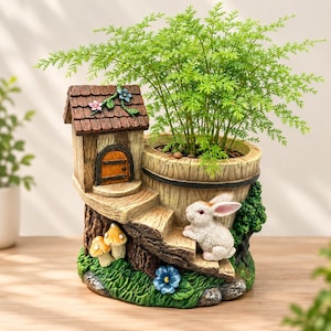Op de afbeelding: Een decoratieve plantenbak van kunsthars in de vorm van een boomstronk met een klein huisje, een trap en een wit konijnenfiguurtje. De plantenbak heeft een bruin dak, een houten deur, twee kleine paddenstoelen en groene bladeren.