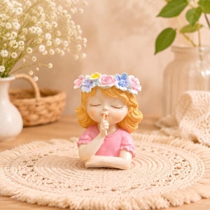May include: A decorative figurine of a young person with wavy blonde hair wearing a crown of pastel pink, blue, and yellow flowers. The figure has closed eyes and a finger held to their lips in a shushing gesture, wearing a pink short-sleeved shirt.
