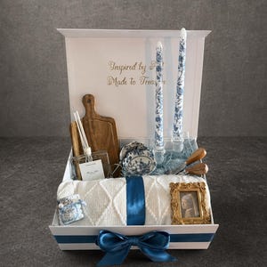 Könnte beinhalten: Eine Geschenkbox mit weißem und blauem Thema, die ein Holzschneidebrett, Kerzen, eine Decke und ein gerahmtes Bild enthält. Die Box ist mit einem blauen Band und einer Schleife verziert. Der Text "Inspired by...Made to Treasure" ist sichtbar.
