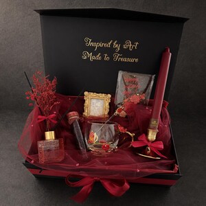 May include: A black gift box with the gold text Inspired by Art Made to Treasure containing a red taper candle in a gold holder, a glass mug with butterfly accents, a small gold frame, a glass diffuser with red dried flowers, and a test tube.