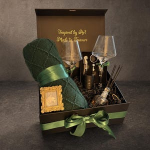 May include: A gift box with a green theme, containing a rolled green blanket with a ribbon, two wine glasses, and other accessories. The box has a green ribbon and the text "Inspired by Art Made to Treasure".