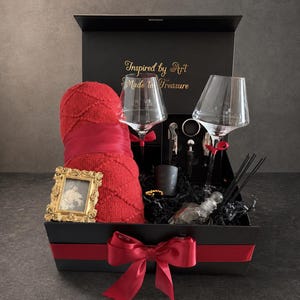 May include: A black gift box with a red ribbon and bow, containing a red towel, two wine glasses, and a small gold picture frame. The box also includes a candle, wine accessories, and a bottle. The text "Inspired by Art Made to Treasure" is on the lid.