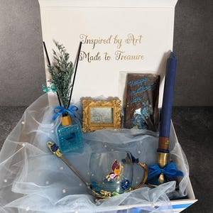 May include: A gift box containing a blue reed diffuser with dried flowers, a small gold ornate frame, a chocolate bar with text, a blue taper candle in a gold holder, and a glass mug with a butterfly design and a matching gold spoon.