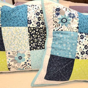 May include: Two square throw pillows with patchwork designs. Each pillow features a variety of floral patterns in shades of blue, white, and green. The pillows have a light blue border and are placed on a white surface.