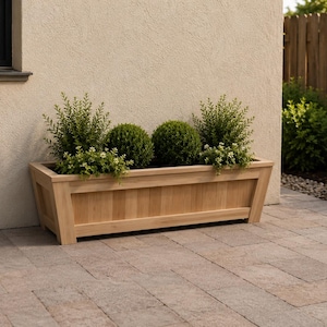 May include: A rectangular wooden planter box with a light brown finish, filled with green plants and shrubs. The planter is placed on a brick patio next to a beige wall and a wooden fence.