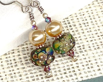 May include: A pair of dangle earrings featuring a combination of beads. Each earring has a silver hook, a champagne-colored bead, a patterned glass bead, and small iridescent crystals. The earrings are displayed on a white surface.