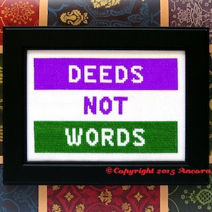 May include: A framed cross-stitch with the text "DEEDS NOT WORDS" in white letters on a purple and green background. The bottom right corner of the frame has the text "Copyright 2015 Ancora Crafts".