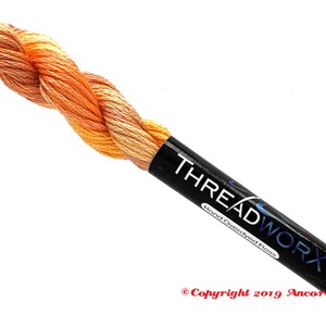 Puede incluir: Un ovillo de hilo de bordar te&ntilde;ido a mano en tonos naranja y amarillo. El hilo est&aacute; envuelto alrededor de una etiqueta negra que dice "THREADWORX Hand Overdyed Floss" y "Copyright 2019 Ancora Crafts".