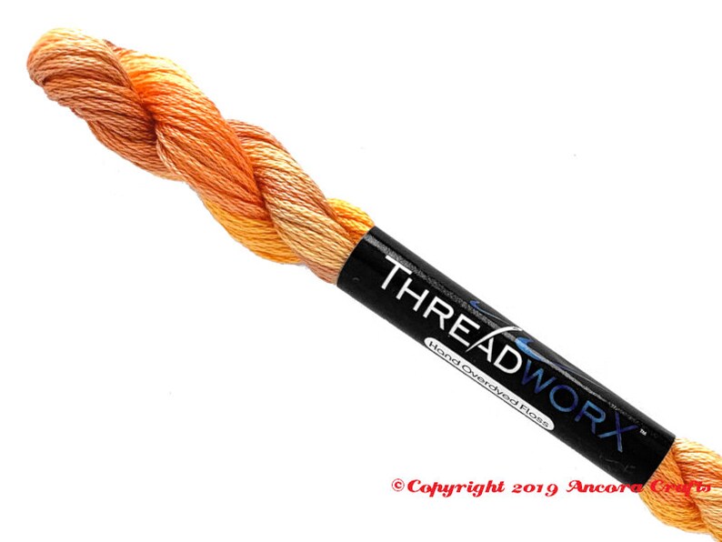 Puede incluir: Un ovillo de hilo de bordar te&ntilde;ido a mano en tonos naranja y amarillo. El hilo est&aacute; envuelto alrededor de una etiqueta negra que dice "THREADWORX Hand Overdyed Floss" y "Copyright 2019 Ancora Crafts".