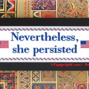 Puede incluir: Bordado blanco de punto de cruz con texto azul que dice "Nevertheless, she persisted" y dos banderas americanas en rojo, blanco y azul. El bordado está enmarcado en negro.