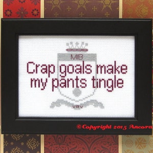 May include: A framed cross-stitch with a white background and burgundy text that reads "Crap goals make my pants tingle". The text is surrounded by a gray and white crest with the letters "MIB" and "VRV" at the top and bottom. The bottom of the frame has the text "Copyright 2015 Ancora Crafts".