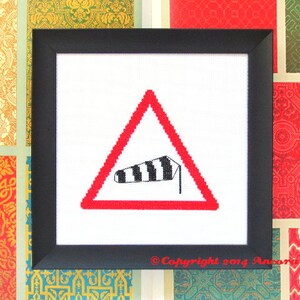 Puede incluir: Un diseño de punto de cruz enmarcado en negro de una señal de advertencia roja y blanca con una manga de viento rayada en blanco y negro que sopla hacia la izquierda. El cartel dice "Copyright 2014 Ancora Designs".