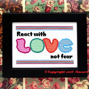 May include: A framed cross-stitch design with the text "React with LOVE not fear" in colorful letters. The design is surrounded by a black frame and a white background.