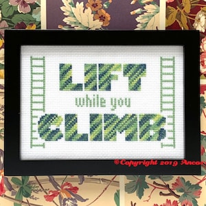May include: A framed cross-stitch design with green and blue stripes that reads "Lift while you climb". The design is framed in black and has a white background.