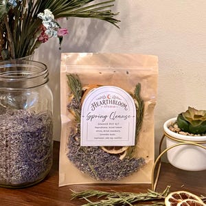 Op de afbeelding: Een doorzichtige plastic zak met een sudderpotset met het label Hearthbloom Studio Spring Cleanse. De set bevat gedroogde citroenschijfjes, rozemarijntakjes en lavendelknoppen. Het etiket vermeldt de ingrediënten: gedroogde citroen, rozemarijn, lavendel.