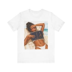 May include: White t-shirt featuring an illustration of a person relaxing on a beach, holding a book over their face. The person is wearing a white bikini. The book has a moon and stars design. The background shows the ocean and sky.
