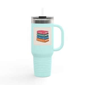 May include: A light blue travel mug with a silver lid and handle. The mug features a graphic of a stack of books with the words "Book", "Hangover", "In", and "Progress". A white straw is inserted into the lid.