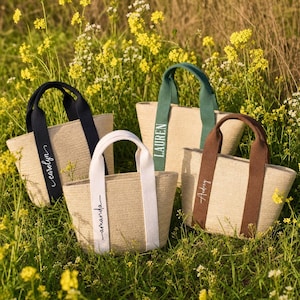 Puede incluir: Cuatro bolsos de paja trenzada con asas de tela resistentes en negro, blanco, verde y marrón. Cada bolso presenta un nombre bordado en una de las asas: Carolyn, Amanda, Lauren y Audrey. Los bolsos tienen un diseño de apertura superior.