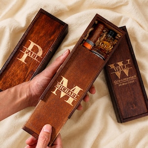 Puede incluir: Tres cajas deslizantes de madera marrón oscuro con grabados personalizados. Una caja está abierta, revelando una pequeña botella de vidrio y un cigarro en su interior. Las tapas muestran los nombres Paul, Michael y Will, con Groomsman 30 May 2024.