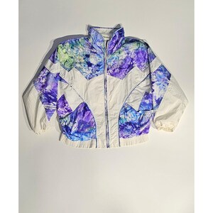 May include: A vintage white and patterned jacket with a high collar and a full zipper. The jacket features a geometric design with purple, blue, and green watercolor-style patterns. The sleeves are long and the jacket has a ribbed hem.