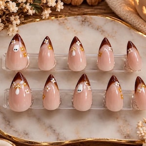 Könnte beinhalten: Ein Set aus zehn mandelförmigen Press-on-Nägeln mit nudefarbener Basis und dunkelbraunen French-Spitzen. Das Design umfasst kleine weiße und gelbe Blumenakzente sowie zwei Nägel, die mit einer kleinen weißen Geisterfigur auf der Spitze verziert sind.