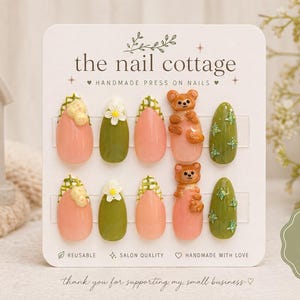 Può includere: Un set di dieci unghie press-on su un cartoncino bianco con la scritta the nail cottage. Le unghie presentano un mix di colori pesca e verde oliva, con decorazioni 3D che includono fiori bianchi, motivi a quadri verdi e orsetti marroni.