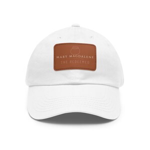 May include: White baseball cap with a curved brim. A brown rectangular patch on the front displays the text "MARY MAGDALENE THE REDEEMED" in a serif font, along with a simple illustration of a pot.