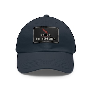 May include: Navy blue baseball cap with a curved brim. The cap features a rectangular patch with the text "RAHAB THE REDEEMED" in white, and a red graphic. The cap is made of a soft, durable fabric.