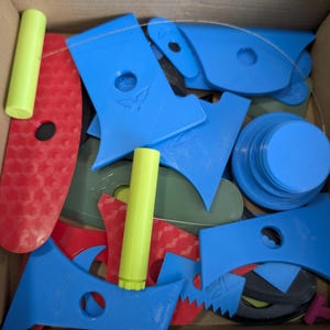 Op de afbeelding: Een verzameling 3D-geprinte gereedschappen in een kartonnen doos, waaronder blauwe, rode en olijfgroene platte schrapers met ronde uitsparingen, twee felgele cilindrische handvatten en een dunne draadlus, allemaal met diverse geometrische vormen.
