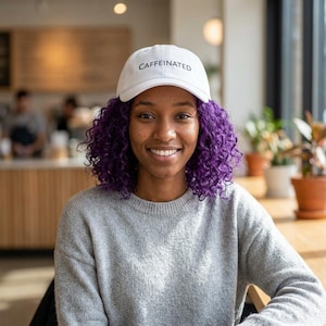May include: A white baseball cap with the word "CAFFEINATED" in black lettering. The person in the image is wearing a gray sweater and has purple curly hair. The background includes a cafe setting with plants and natural light.