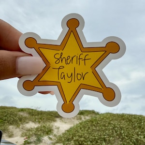 May include: A yellow six-pointed star-shaped sticker with a white border. The sticker features the text Sheriff Taylor written in black handwritten-style lettering across the center. The design mimics a traditional law enforcement badge shape.