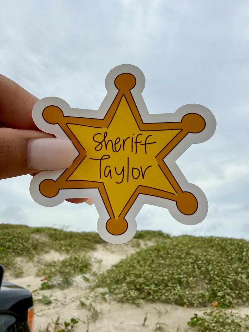 May include: A yellow six-pointed star-shaped sticker with a white border. The sticker features the text Sheriff Taylor written in black handwritten-style lettering across the center. The design mimics a traditional law enforcement badge shape.