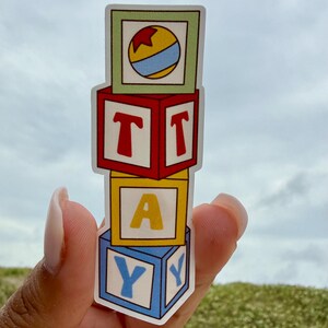 May include: A vertical sticker featuring four stacked square toy blocks. The top block is light green with a yellow, red, and blue ball. The second block is red with the letter T. The third block is yellow with the letter A. The bottom block is blue with the letter Y.