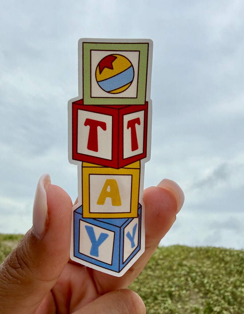 May include: A vertical sticker featuring four stacked square toy blocks. The top block is light green with a yellow, red, and blue ball. The second block is red with the letter T. The third block is yellow with the letter A. The bottom block is blue with the letter Y.