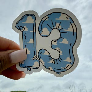 May include: A die-cut sticker shaped like the number 13, designed to look like a foil balloon. The sticker features a light blue sky background with white, fluffy clouds and black outlines that define the shape and the balloon's crinkled texture.