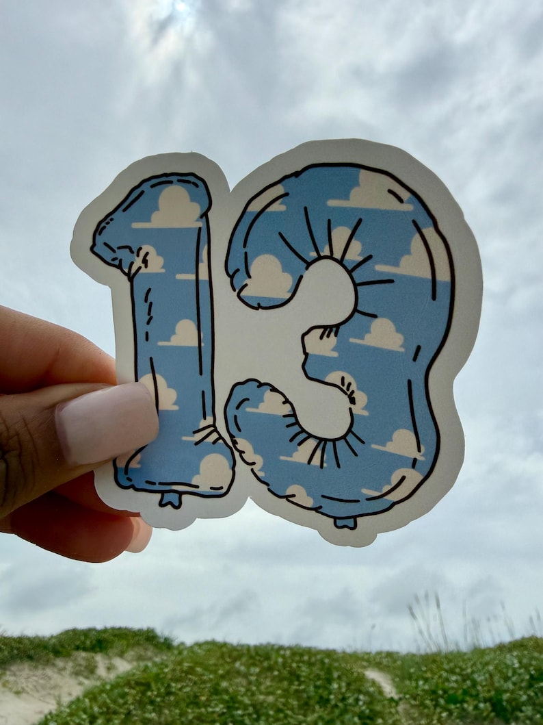 May include: A die-cut sticker shaped like the number 13, designed to look like a foil balloon. The sticker features a light blue sky background with white, fluffy clouds and black outlines that define the shape and the balloon's crinkled texture.