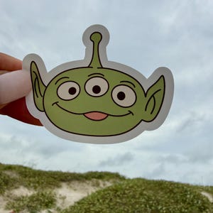 May include: A die-cut vinyl sticker featuring a cartoon alien character with light green skin, three large eyes, pointed ears, and a small antenna on top of its head. The character has a wide, friendly smile with a visible pink tongue.