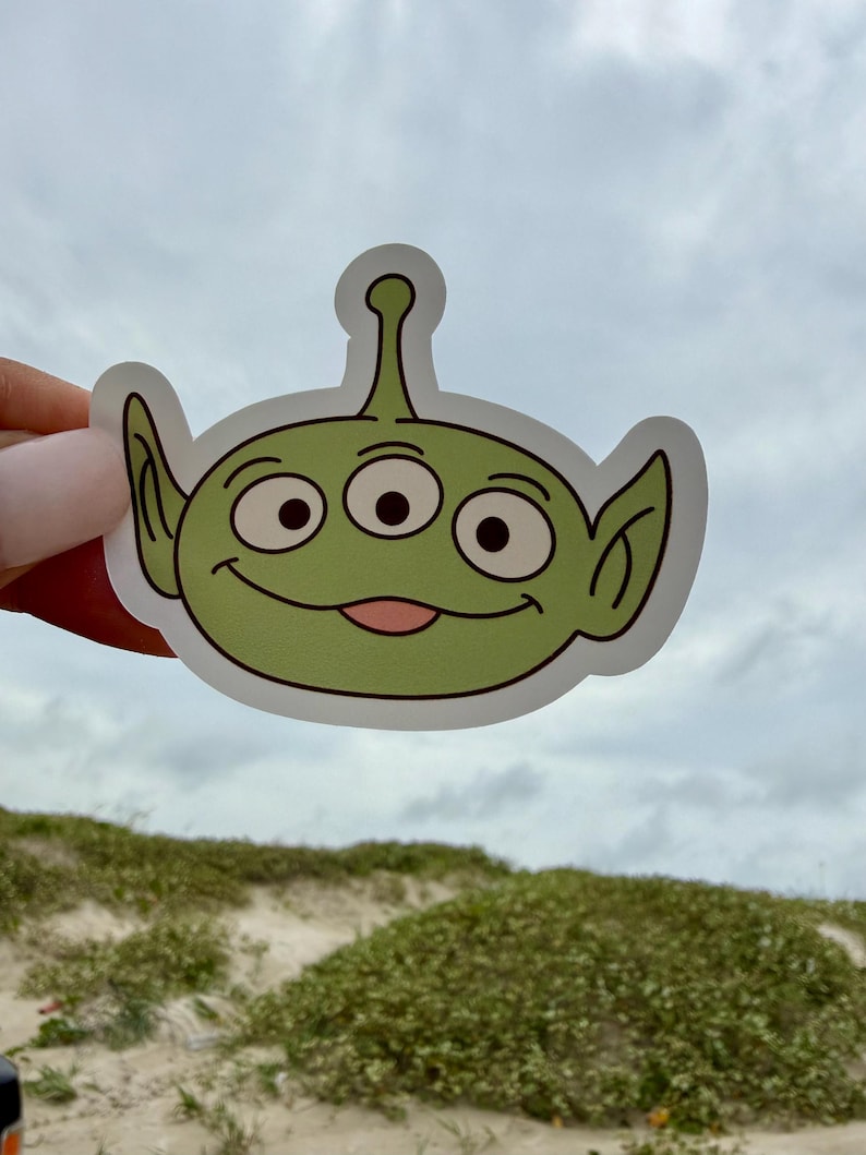 May include: A die-cut vinyl sticker featuring a cartoon alien character with light green skin, three large eyes, pointed ears, and a small antenna on top of its head. The character has a wide, friendly smile with a visible pink tongue.