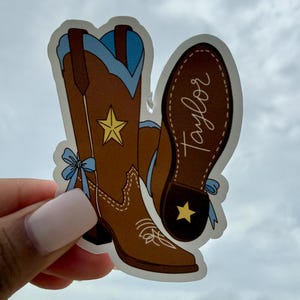 May include: A die-cut sticker featuring two brown cowboy boots with light blue accents, a yellow star on the shaft, and a light blue bow on the side. One boot shows the sole with the name Taylor written in white script and a yellow star on the heel.