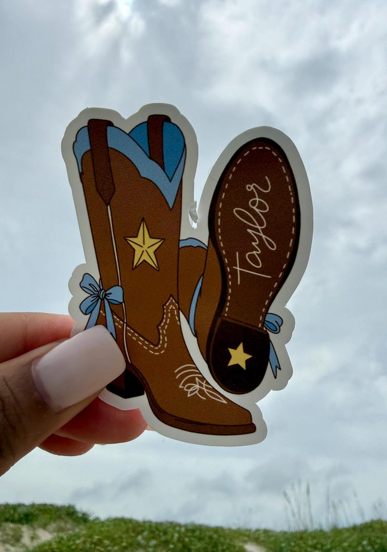 May include: A die-cut sticker featuring two brown cowboy boots with light blue accents, a yellow star on the shaft, and a light blue bow on the side. One boot shows the sole with the name Taylor written in white script and a yellow star on the heel.