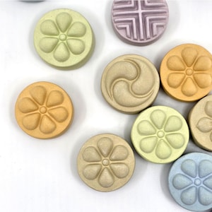 May include: A collection of round, decorative ceramic pieces in various pastel colors. Some feature floral designs, while others have geometric or abstract patterns. The pieces are arranged on a white surface, showcasing their different shapes and hues.