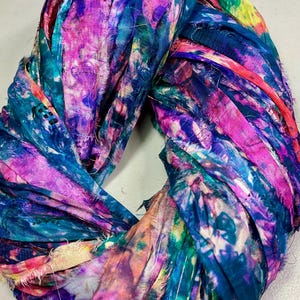 May include: A vibrant, hand-dyed textile in a circular arrangement. The fabric displays a rich palette of colors, including blues, purples, pinks, and greens. The material appears to be soft and flowing, with a slightly frayed edge, suggesting a scarf or wrap.