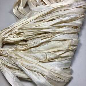 May include: Cream-colored silk ribbon in a tangled bundle. The ribbon has a slightly textured appearance with frayed edges, suggesting a handmade or vintage quality. The color is a soft, off-white, with subtle variations in tone.